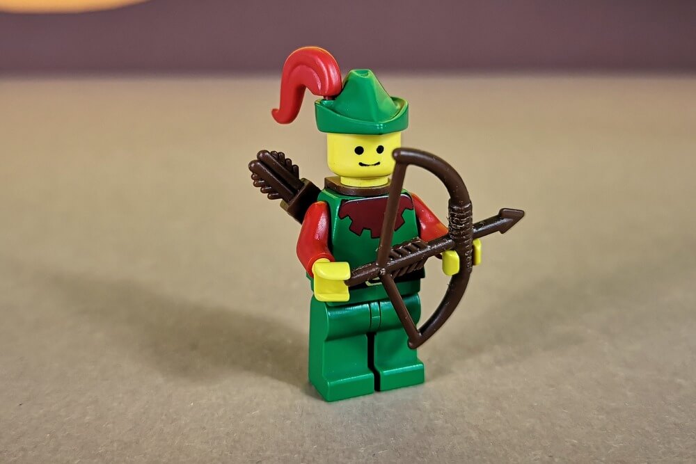 Bogenschütze in grüner Kleidung der Forestmen mit roten Armen und roter Feder am grünen Hut. In der Hand hat er einen Bogen.