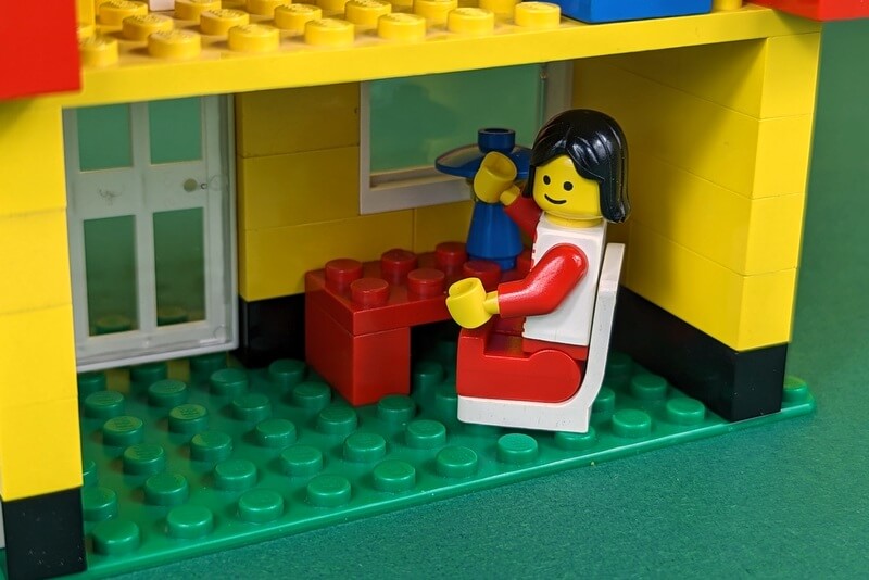 Eine Lego-Figur sitzt an einem Schreibtisch in einem Lego-Haus und schaut lächeln in die Kamera.