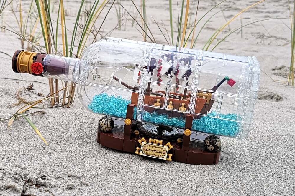 Wenn man alle Bausteine zusammengesteckt hat, steht das fertige Flaschenschiff vor einem. Zu sehen ist ein Segelschiff aus LEGO-Teilen in einer Flasche, die aus transparenten LEGO-Panels gebaut ist. Im Bild steht das fertige Modell am Strand im Sand. 