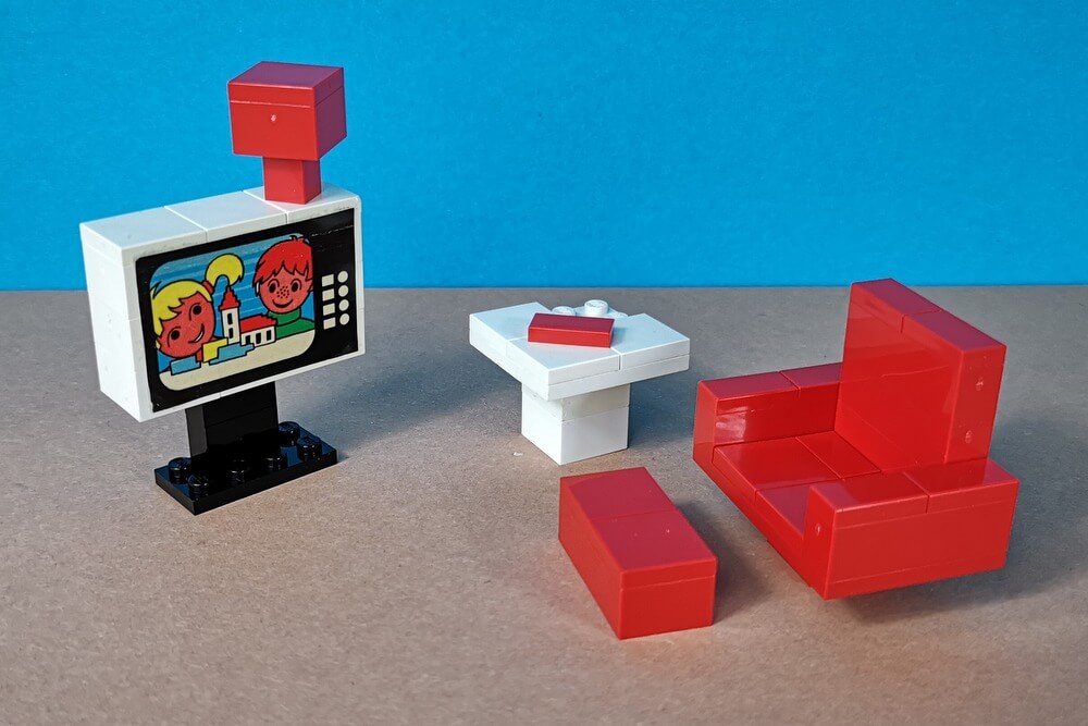 Zu sehen ist ein Fernseher im klassischen Look mit einem bequemen roten Sessel, einem kleinen Tisch und einer roten Fußbank. Alles Möbel sind aus Lego-Steinen gebaut.