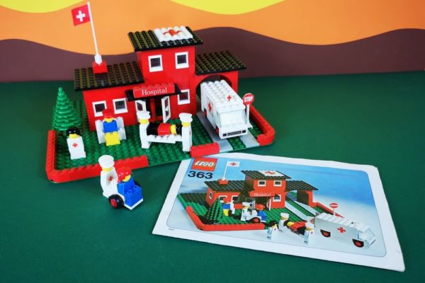 LEGO-Set 363 vor typischem Fotohintergrund der 70er-Jahre, der aus Pappe besteht und Wiese, Hügel und Himmel zeigt. Set 363 ist ein Krankenhaus in der interessanten Farbe Rot. Im Vordergrund liegt die Bauanleitung von 1975.