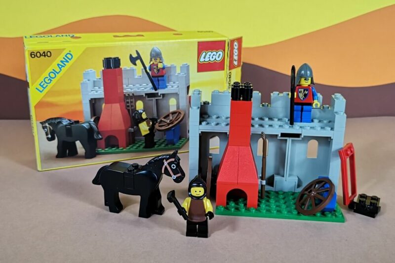 Vor einem schönen Bilderbuchhintergrund steht Lego-Set 6040 in aufgebautem Zustand mit der sehr seltenen Original-Verpackung. Die kleine Schmiede kommt mit einem Ritter, einem Schmied und einem schwarzen Pferd.