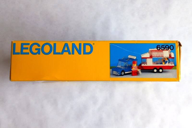 Die Oberseite ist gelb mit dem typischen blauen Legoland-Schriftzug der 80er-Jahre und einem kleinen Bild vom Wohnwagen mit Auto.