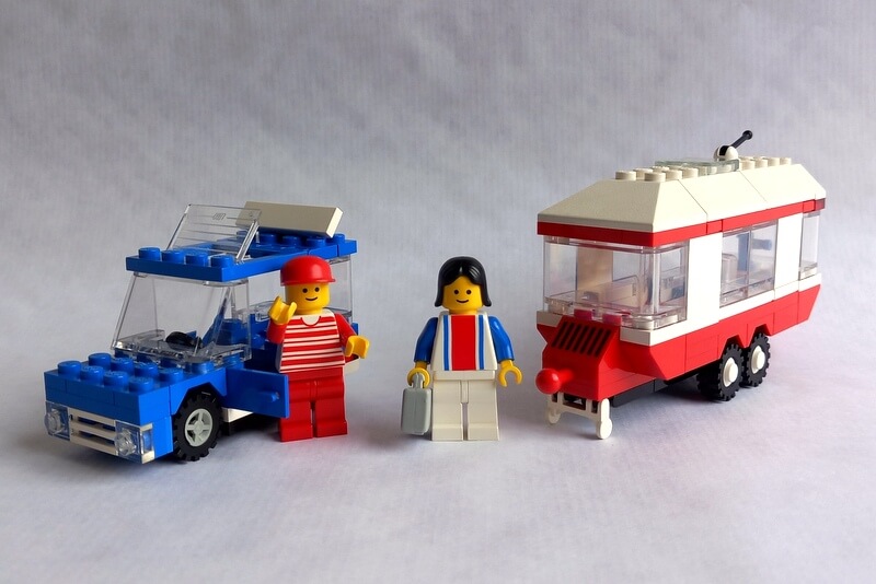 Ein winkendes Lego-Männchen und eine Lego-Frau vor ihrem Auto und Wohnwagen kurz vor der Reise in den Urlaub.