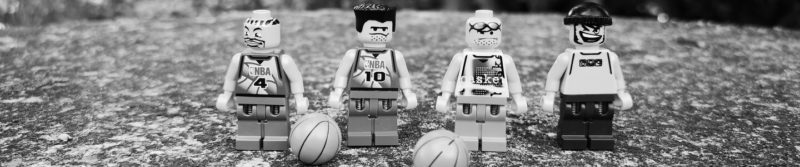 Das Bild zeigt vier Minifiguren aus der LEGO-Basketball-Serie und zwei Basketbälle. Die Figuren haben Federn in den Beinen und können so Bälle schnippend werfen.