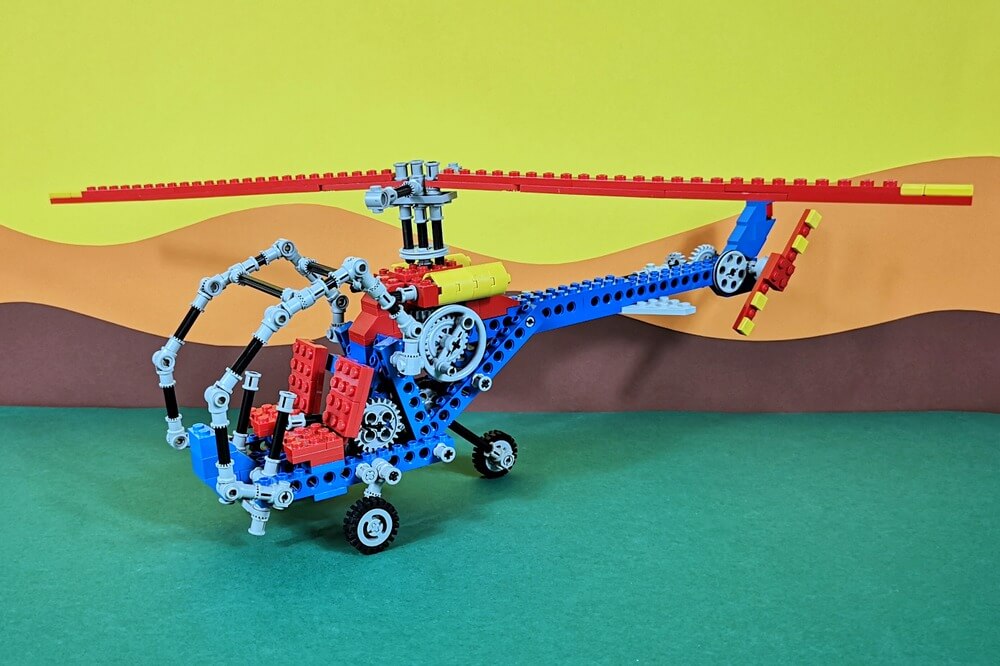 Ein klassischer Lego-Hubschrauber aus Technik-Bausteinen in den Farben blau, rot, grau, schwarz und etwas gelb. Er wurde vor buntem Hintergrund fotografiert, der aussieht wie Wiese, Berge und Himmel mit Sonne.