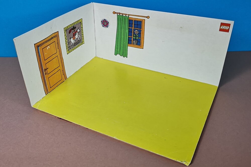 Eine Kulisse aus Pappe für die Sets der Home-Maker-Serie. Der Fußboden ist gelb, die Wände sind weiß mit Bildern und einem Fenster und es gibt sogar eine Tür, die aus Pappe ausgeschnitten ist.