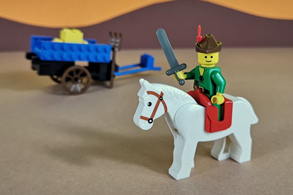 Ein roter Sattel macht das weiße Pferd zu einem Reitpferd. Der Heuwagen steht abgekoppelt im Hintergrund geparkt.