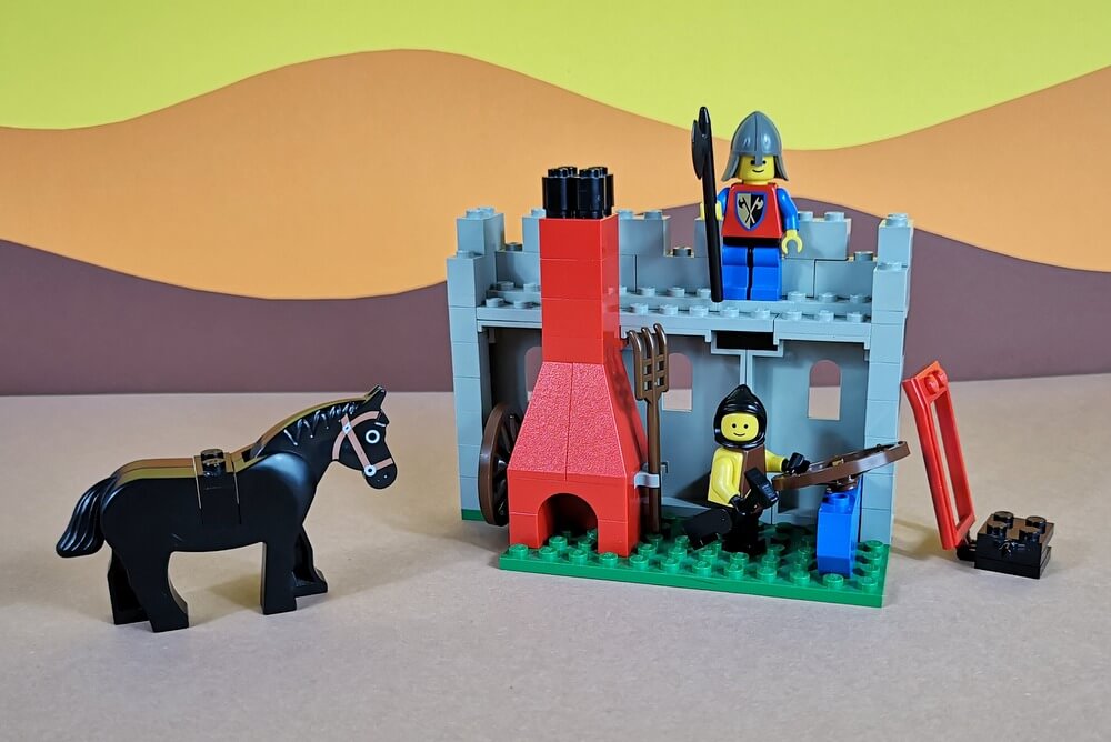 Die Schmiede ist aus grauen Lego-Steinen gebaut, hat einen roten Schmiedeofen und einen blauen Amboss. Ein schwarzes Pferd, ein Schmied und eine Wache sind auch mit dabei.