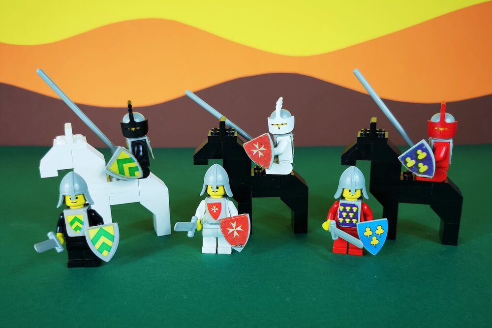 Drei Turnier-Ritter auf Pferden und Landsknechte mit Schwertern. Es sind ingesamt drei verschiedene Wappen auf den Schilden zu sehen.